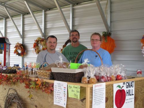 Three men stand behind a wooden stall filled with baskets of colorful treats, surrounded by autumn decorations.