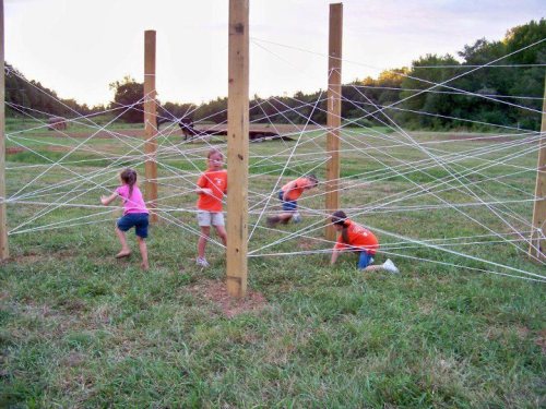 Children navigate a web of ropes between wooden posts in a grassy field during an outdoor activity.