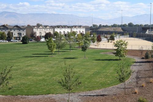 A grassy park with trees, surrounded by residential buildings and a tennis court in the background under a cloudy sky.