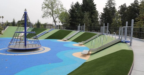 A colorful playground featuring slides, a climbing structure, and grassy areas surrounded by trees.