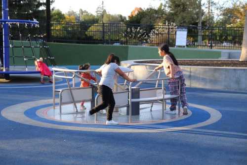 Children playing on a spinning playground structure in a park, with trees and a playground in the background.