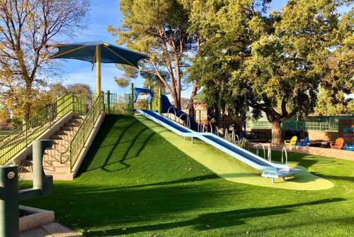 A playground featuring a green hill with two slides, stairs, and shaded areas under trees.