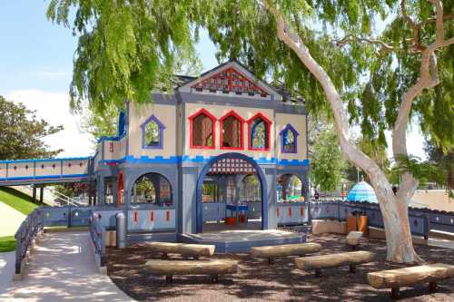 Colorful playground structure with arches, surrounded by trees and benches, set in a sunny outdoor area.