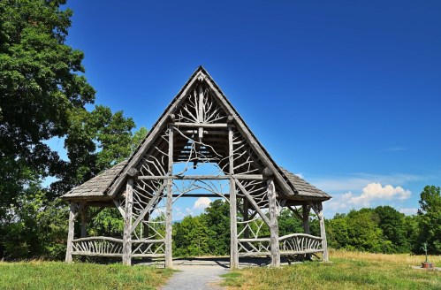 A wooden pavilion with a unique design stands in a grassy field under a clear blue sky.