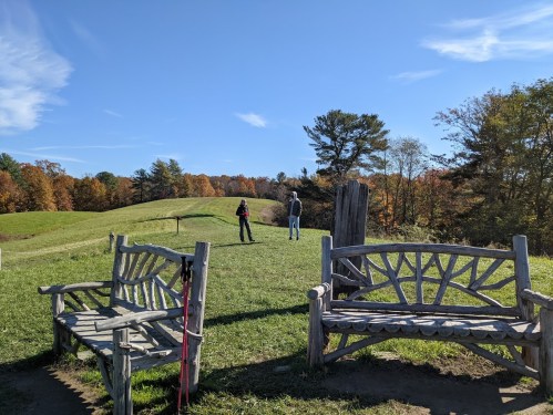 Two people stand in a grassy field with autumn trees in the background, framed by rustic wooden benches.