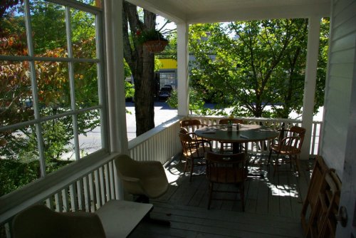 A sunny porch with a round table and wooden chairs, surrounded by greenery and large windows.