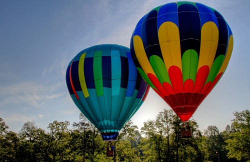 Two colorful hot air balloons in the sky, one blue and green striped, the other red and yellow, surrounded by trees.