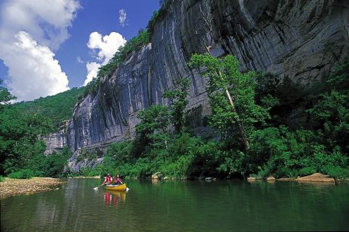 Two people kayaking on a calm river surrounded by lush greenery and towering cliffs under a blue sky with clouds.