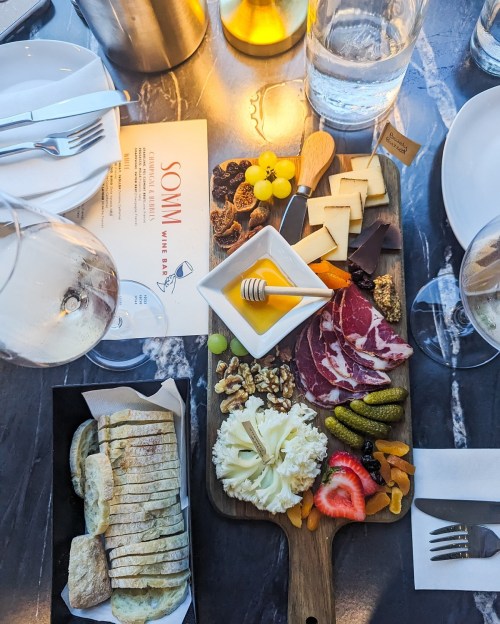 A charcuterie board with cheeses, meats, fruits, nuts, and honey, accompanied by wine glasses and a menu on a table.