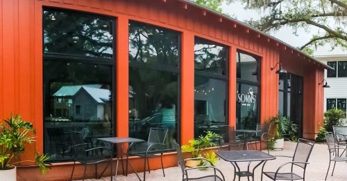 Exterior of a modern restaurant with large windows, red siding, and outdoor seating surrounded by greenery.
