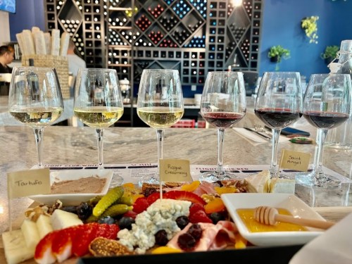A wine tasting setup with six glasses of wine and a charcuterie board featuring cheeses, meats, fruits, and honey.