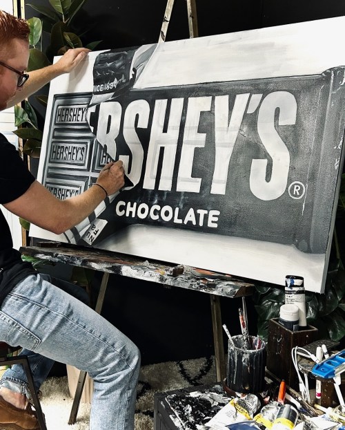 An artist paints a black-and-white depiction of a Hershey's chocolate bar on a canvas, surrounded by art supplies.