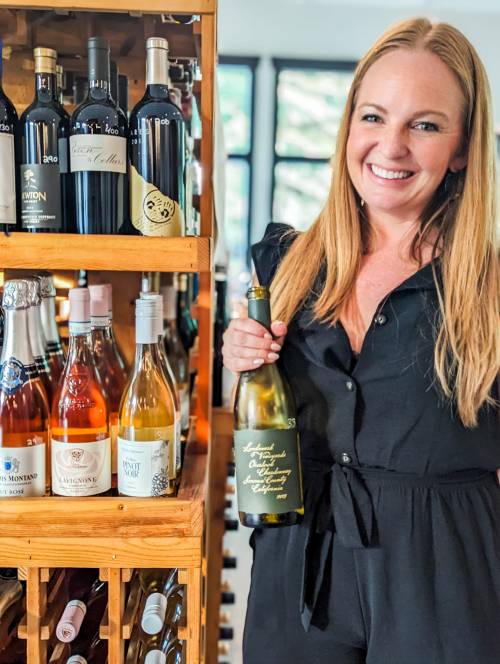 A woman in a black outfit smiles while holding a bottle of wine next to a wooden wine rack filled with various bottles.
