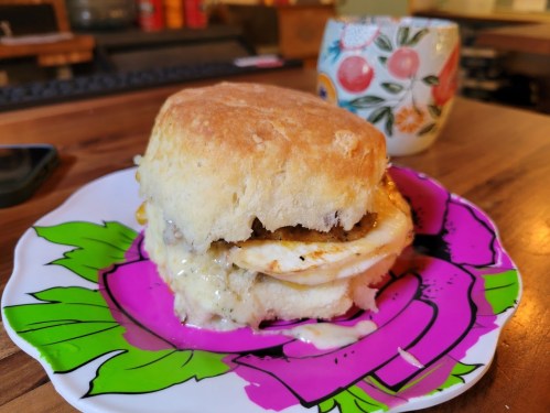 A fluffy biscuit sandwich filled with egg and sausage, served on a colorful floral plate with a mug in the background.