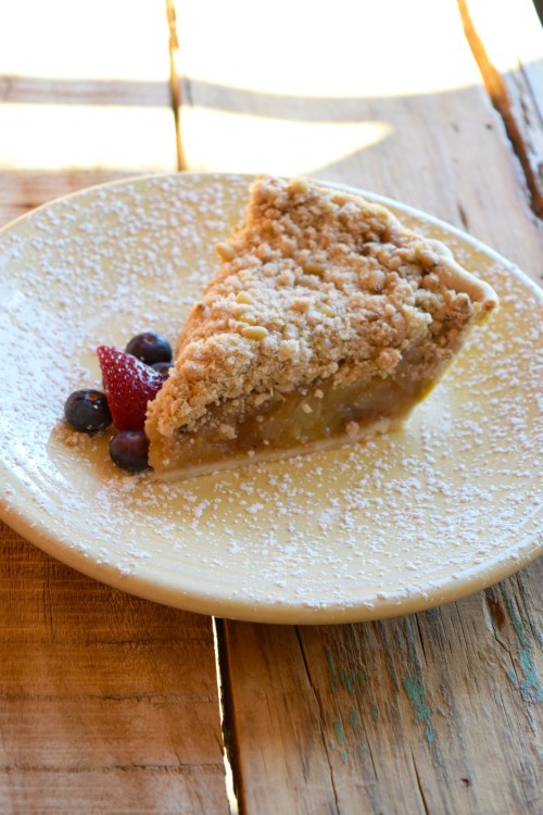 A slice of apple crumble pie on a plate, garnished with strawberries and blueberries, dusted with powdered sugar.