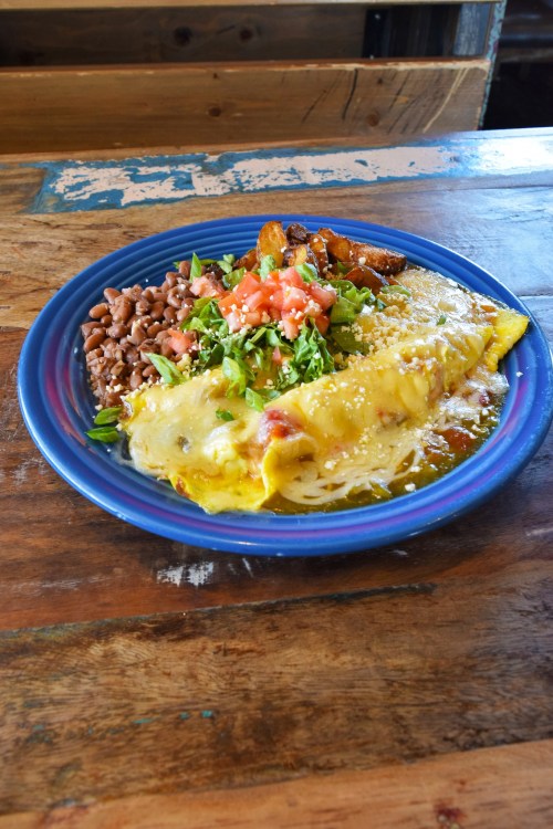 A colorful plate featuring a cheese omelette, beans, plantains, diced tomatoes, and chopped greens on a wooden table.