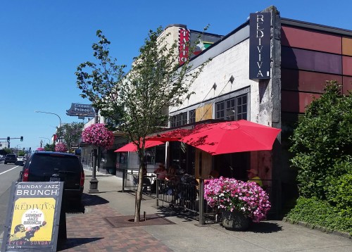 A restaurant exterior with red umbrellas, flower planters, and a sign for Sunday jazz brunch.
