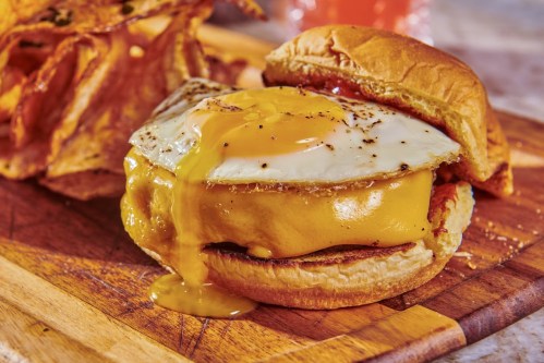 A delicious sandwich with a melted cheese patty, a fried egg, and crispy potato chips on a wooden board.