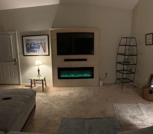 Cozy living room with a TV above an electric fireplace, a lamp, and a glass shelf, all on soft carpet.
