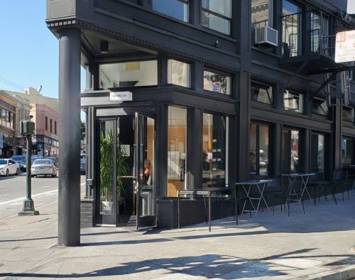 A modern storefront with large windows, featuring a door and outdoor seating, located on a city street.