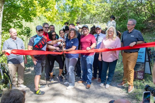 A group of people gathers to cut a red ribbon at a trail opening event, celebrating the new community path.