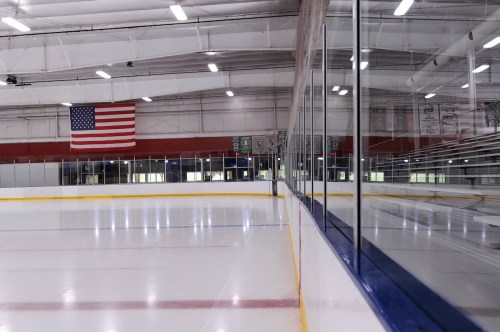 An empty ice rink with a glass barrier and an American flag hanging in the background.