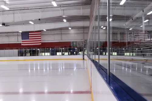 An empty ice rink with a glass barrier and an American flag hanging in the background.