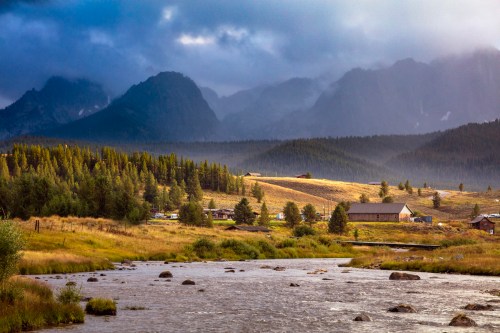 A serene landscape featuring a river, rolling hills, and mountains under a cloudy sky, with a few buildings in the distance.