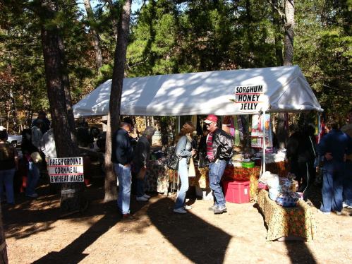 A bustling outdoor market with a tent selling sorghum honey jelly and fresh ground cornmeal, surrounded by trees.