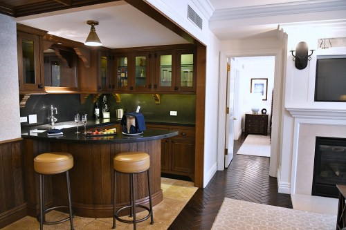 A cozy hotel suite featuring a wooden bar area with stools, a fireplace, and a hallway leading to another room.