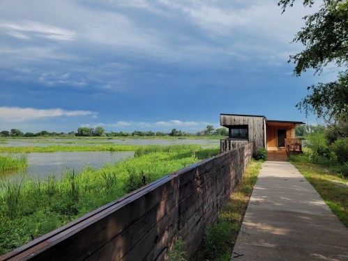 A pathway leads to a wooden building by a serene wetland, with lush greenery and a cloudy sky in the background.
