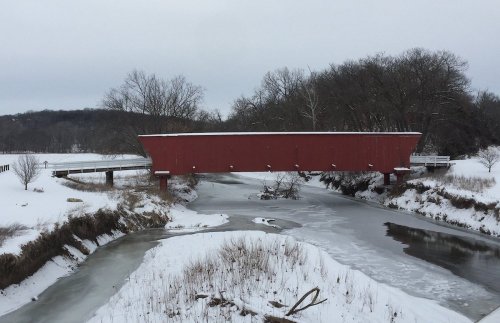 A red covered bridge spans a snowy landscape, with a frozen river and bare trees in the background.