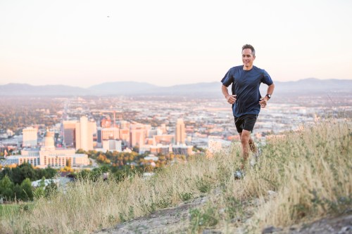 A man runs on a hillside with a city skyline in the background during sunset.