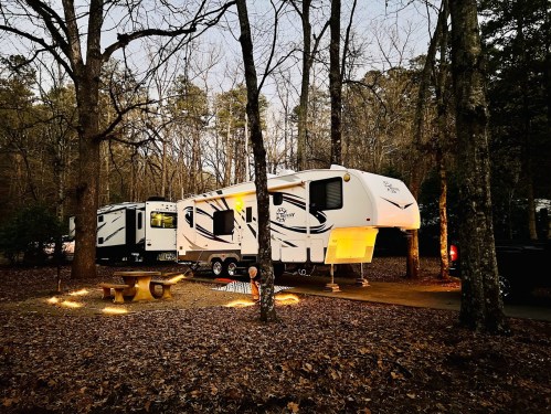 A large RV parked in a wooded area, surrounded by fallen leaves and illuminated by soft outdoor lights at dusk.