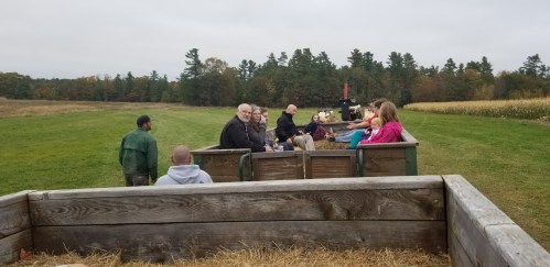 A group of people sitting on a hayride in a field, with trees and a tractor in the background on a cloudy day.
