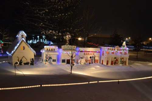 A snowy scene at night featuring colorful holiday lights on miniature buildings, including a church, garage, and hotel.