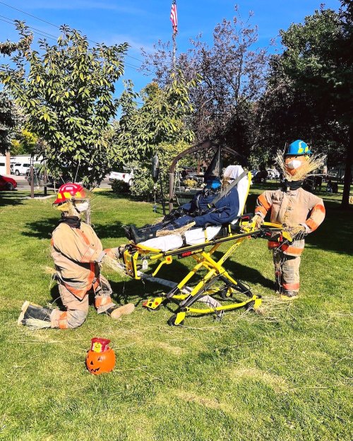 Two scarecrows dressed as firefighters assist a figure on a stretcher in a grassy area, with a pumpkin nearby.