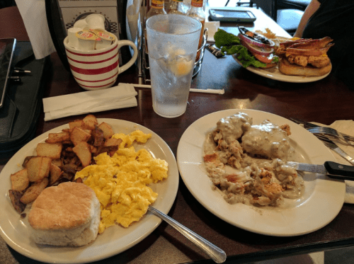 A plate with scrambled eggs, potatoes, a biscuit, and a dish with gravy and meat, alongside a glass of water.