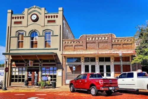 Historic buildings with a clock tower, storefronts, and parked red and white trucks on a sunny day.