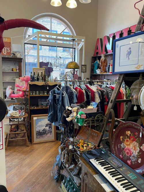 A cozy vintage shop filled with clothing, decor, and eclectic items, featuring a bright window and wooden floors.