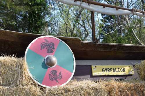 A colorful shield with a green and red design rests against a wooden boat, with a sign reading "GYRFALCON" nearby.