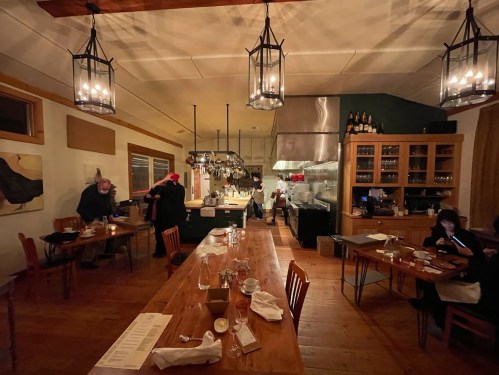 Cozy restaurant interior with wooden tables, hanging lights, and an open kitchen; diners enjoying their meals.