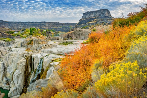 A scenic view of a waterfall surrounded by colorful autumn foliage and rocky cliffs under a cloudy sky.