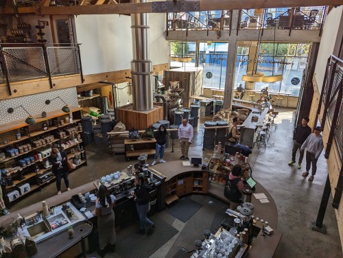 A spacious coffee shop with wooden decor, featuring a large counter, customers, and baristas at work.