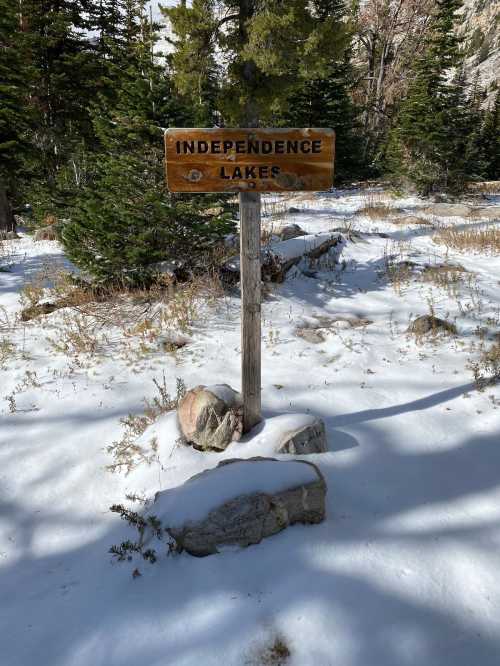 A wooden sign reading "Independence Lakes" stands in a snowy landscape surrounded by evergreen trees.