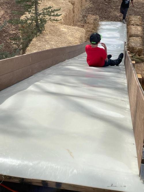 A child in a red shirt sits on a slick slide, ready to go down, with hay bales lining the sides.