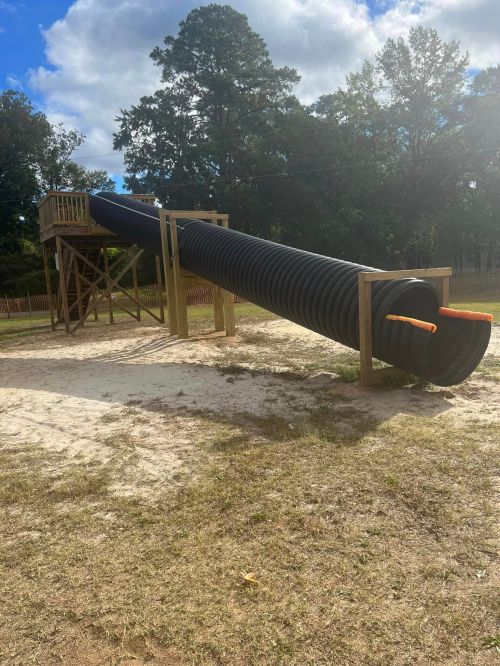 A large black tube slide on a wooden structure in a playground, surrounded by grass and trees under a cloudy sky.