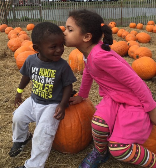 A girl kisses a boy on the cheek while sitting on a pumpkin in a pumpkin patch, surrounded by many pumpkins.
