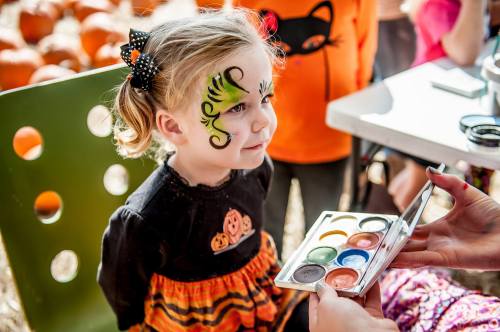 A young girl with a green face painting of a cat sits patiently while someone holds a palette of face paints.