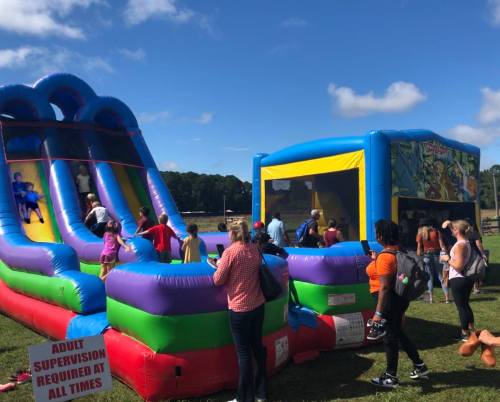 A colorful inflatable bounce house and slide with children playing, set in a grassy field under a blue sky.
